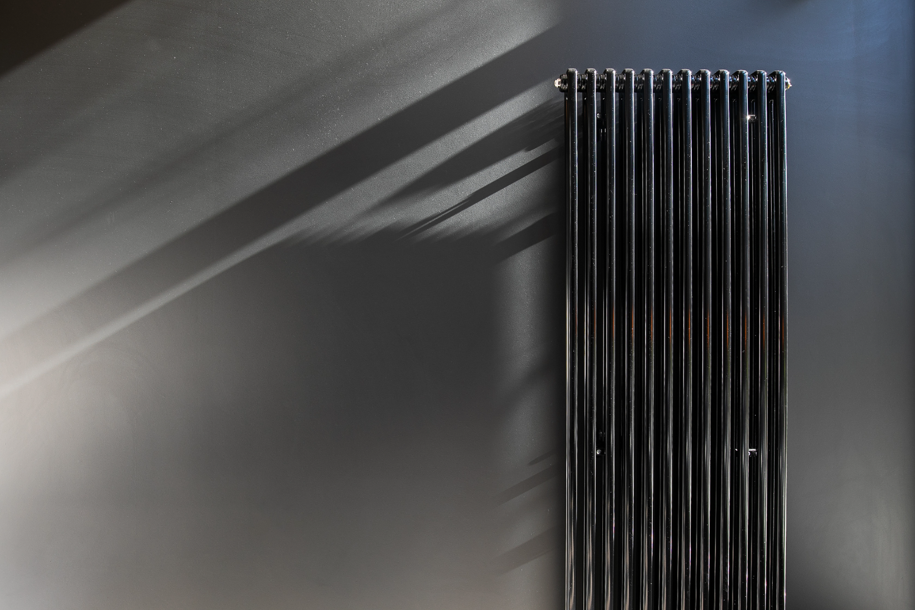  Black Vertical Radiator Mounted Onto A Black Wall With Light Shining In From Window